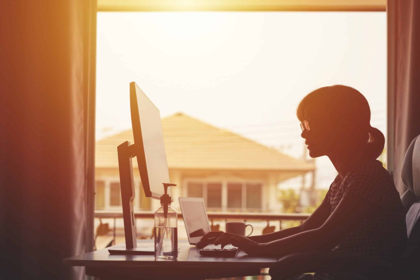 A silhouette of a young women working from home, the sun is setting through her window