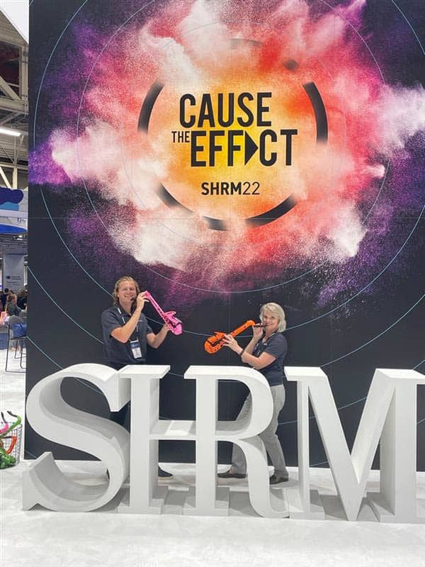 Two DISHER team members pose with fake saxophones in front of a big SHRM 2022 conference sign