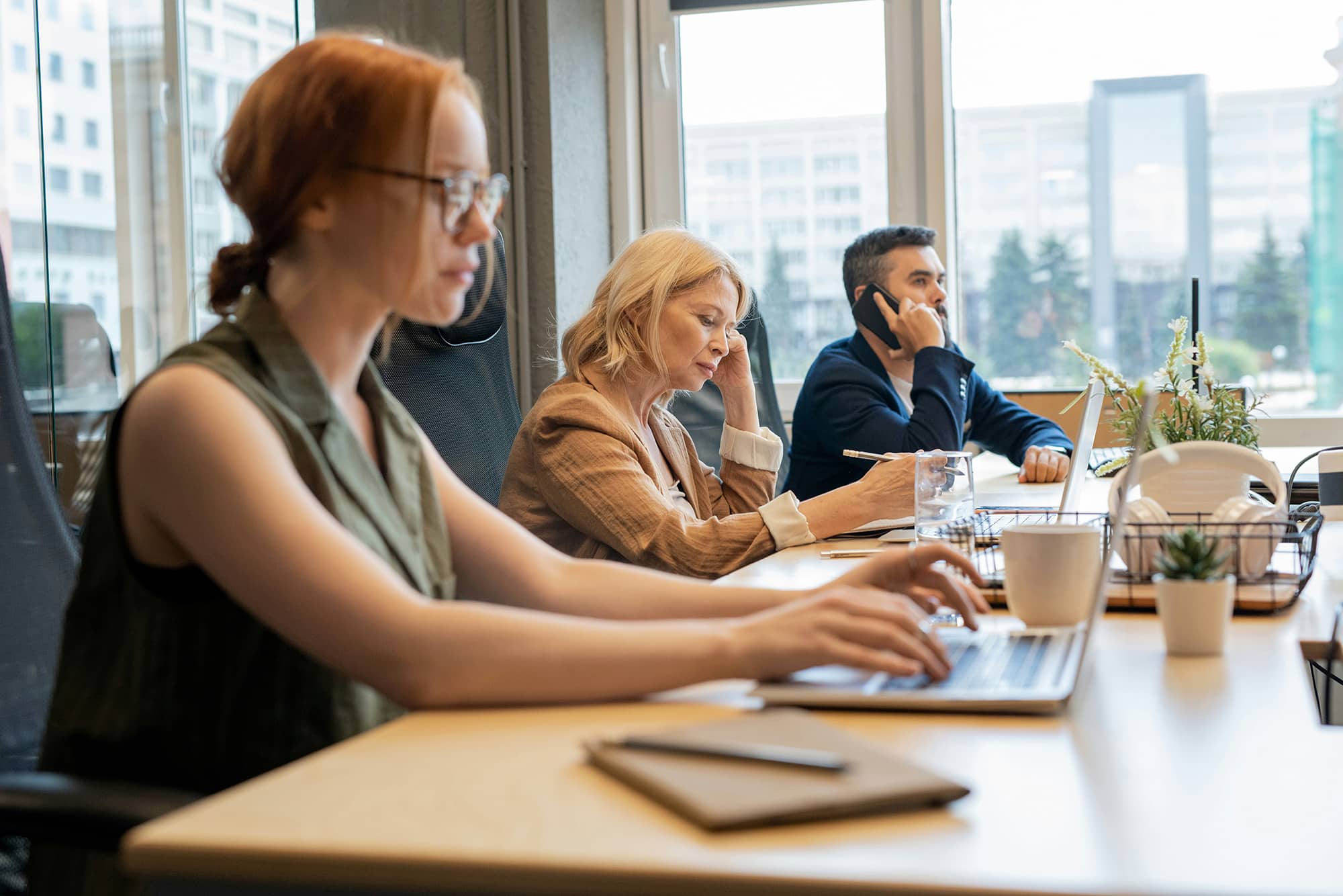 Three coworkers working at a shared desk with laptops, two talking on phones.