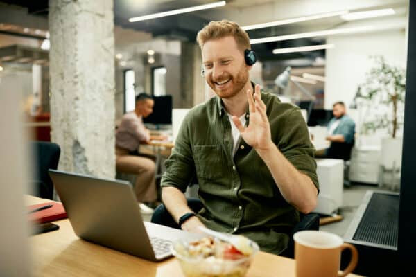 Recruiter on a video call in an open office, waving at a laptop while wearing a headset
