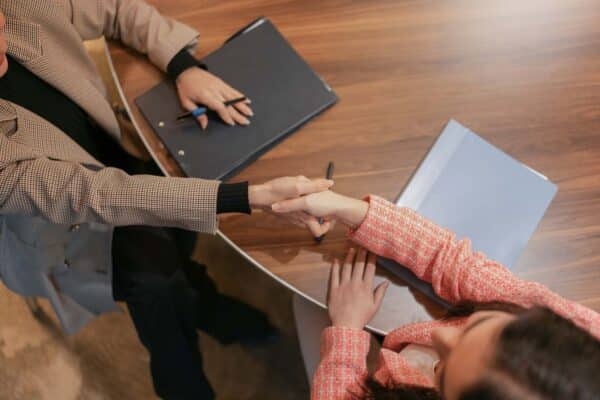 Two professionals shaking hands across a wooden conference table with folders and pens nearby